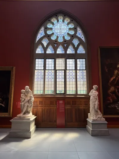 A museum gallery with deep red walls, a tall pointed-arch window of leaded glass, and two white marble sculptures on pedestals.